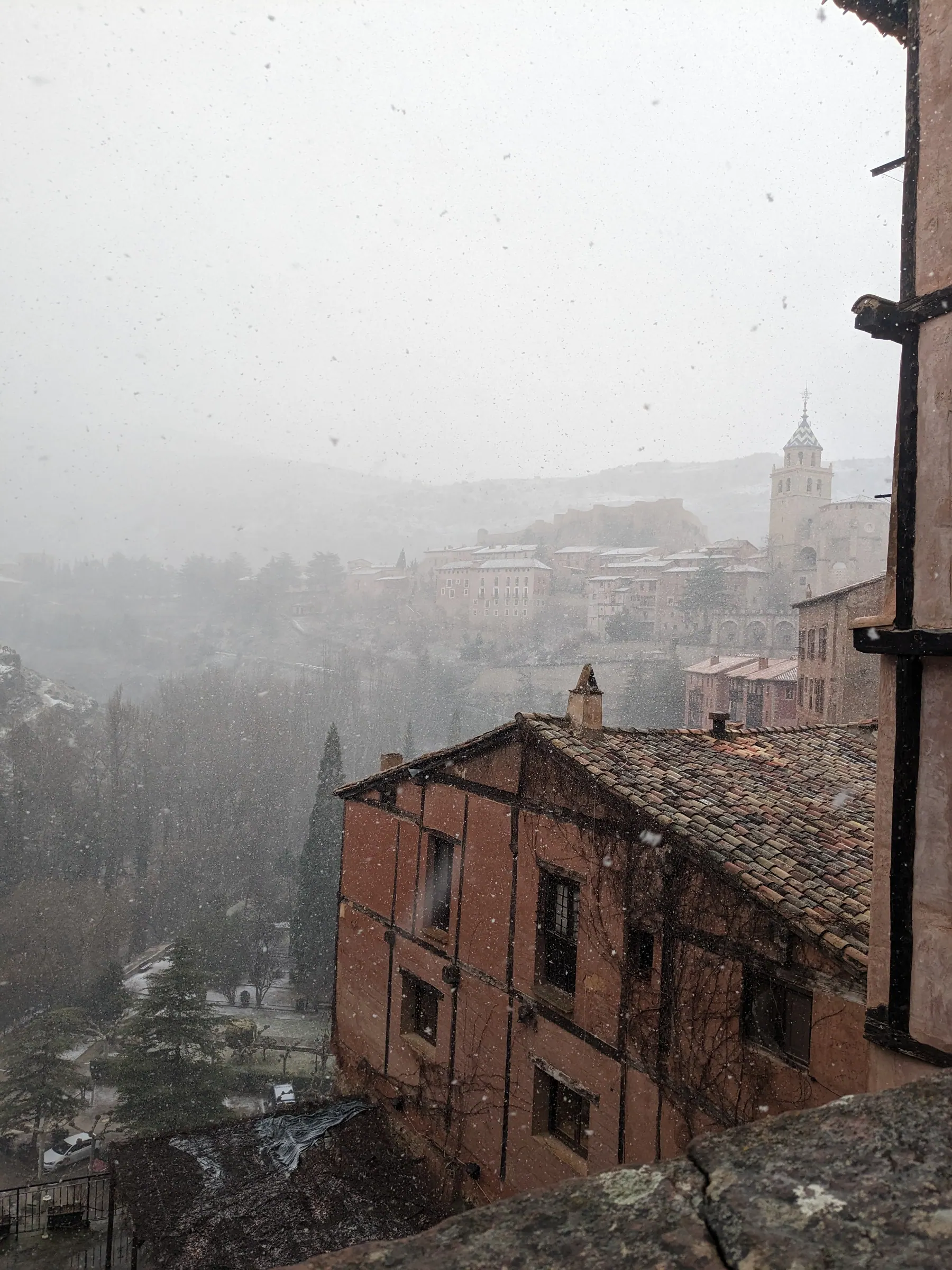 Active snowfall falling over the tiled rooftops and sandstone houses of Albarracín towards the cathedral tower