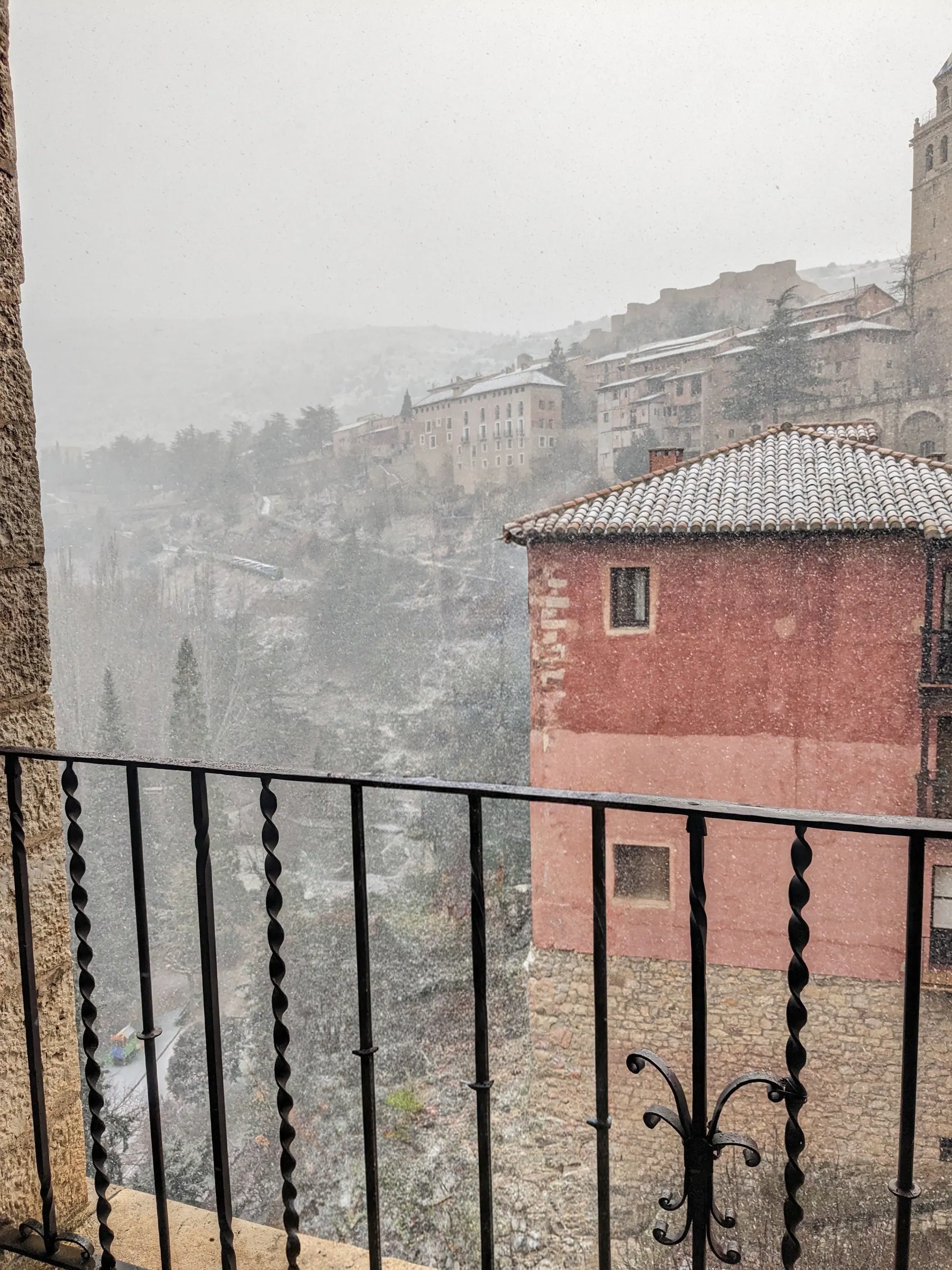 View through the iron balcony railing of Plaza Mayor overlooking snow falling on the rooftops of Albarracín