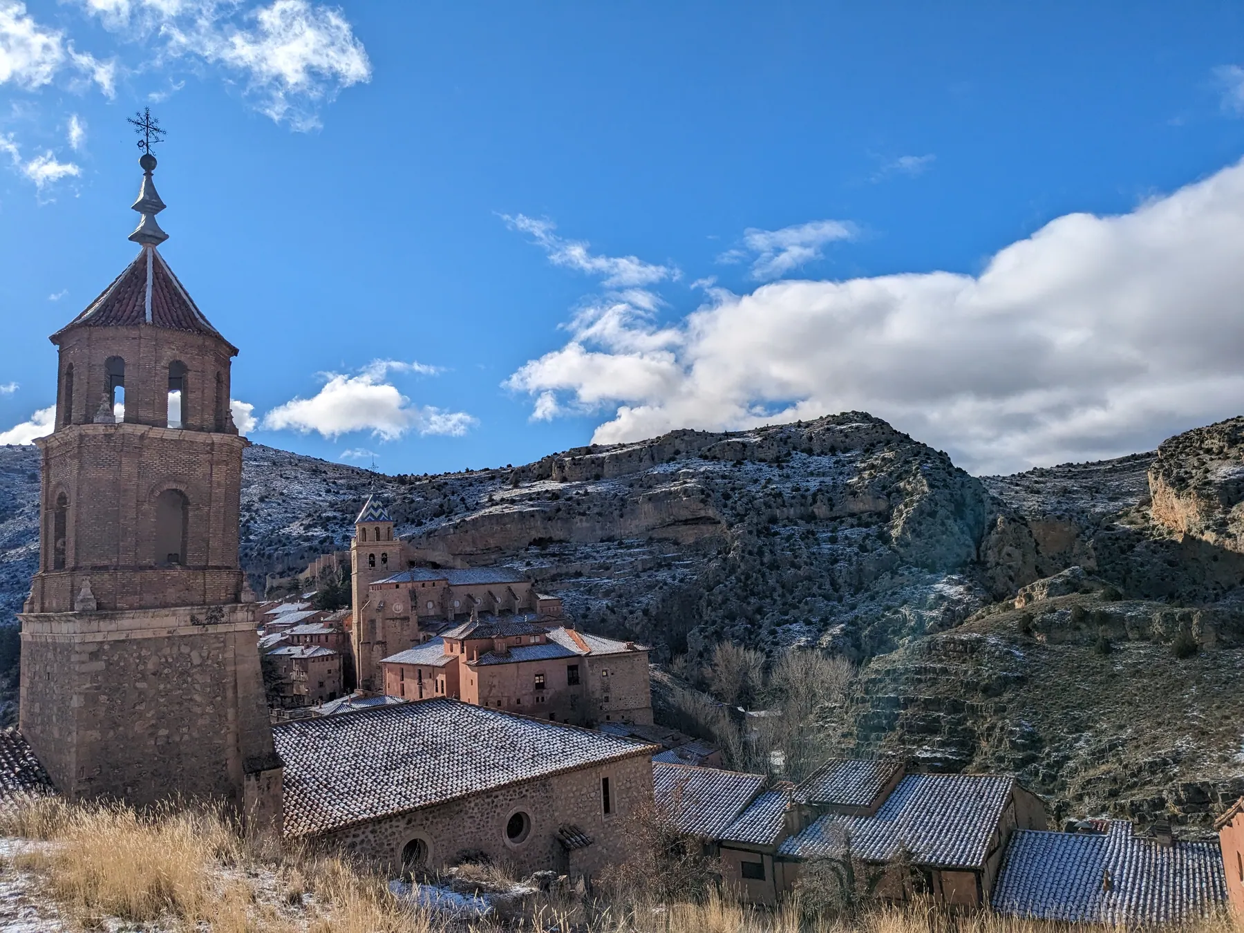 The bell tower of the Iglesia de Santiago Apóstol in the foreground and the Cathedral of Albarracín further in the distance, rising over snow-dusted terracotta rooftops against winter mountain cliffs