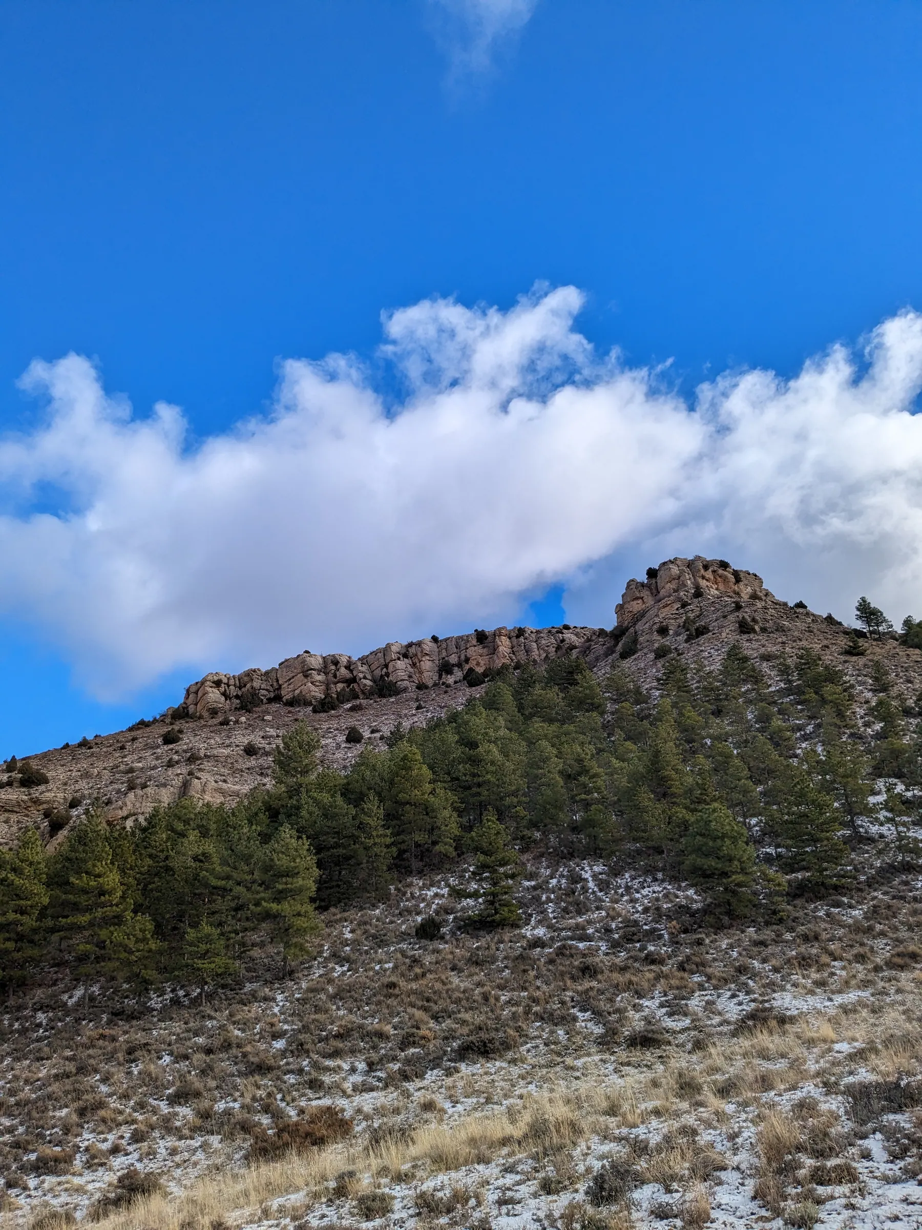 Pine-covered rocky ridge dusted with snow under blue sky and clouds viewed from Mirador de Albarracín