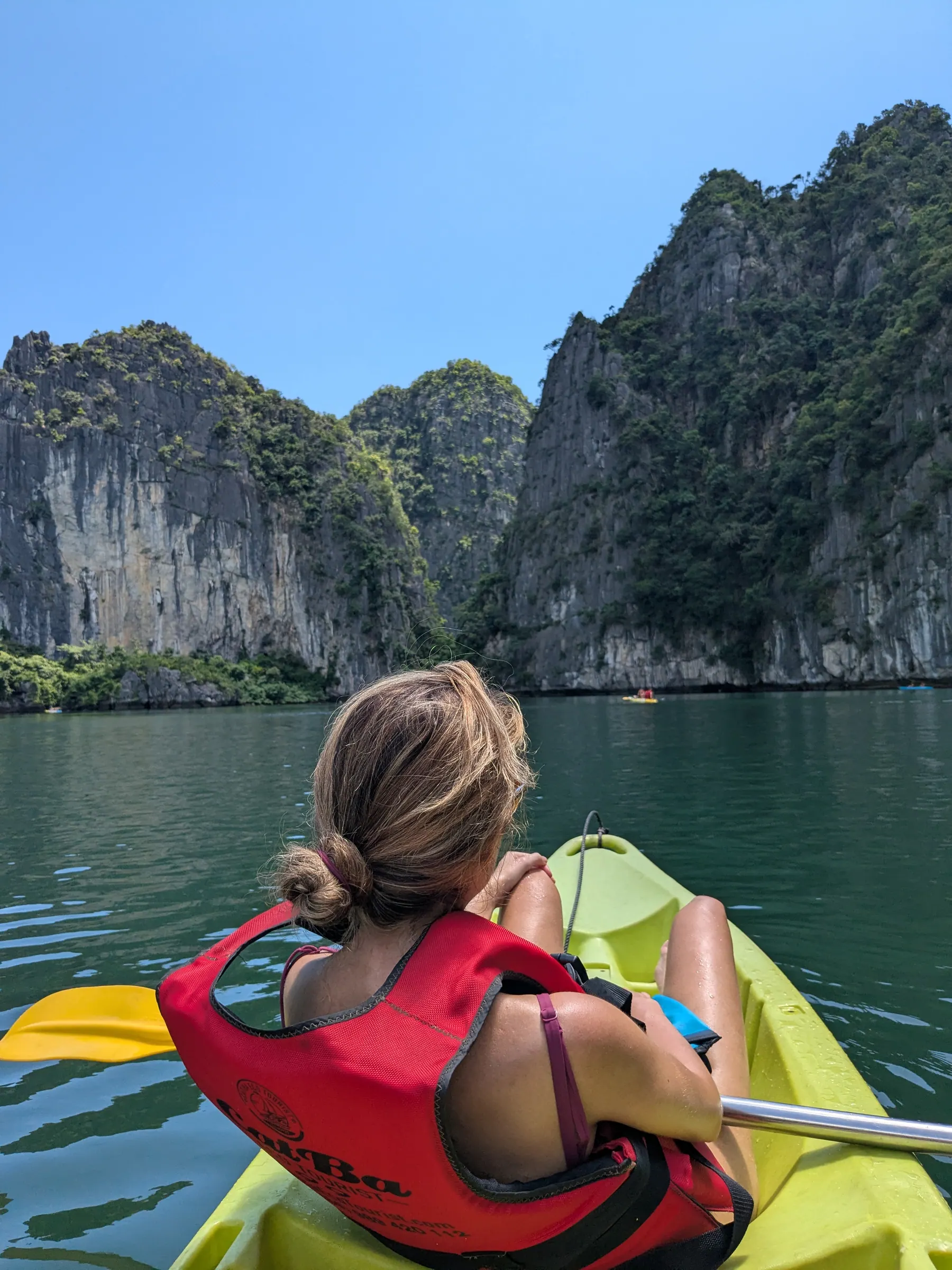 Zane paddling a yellow kayak with a red life jacket across calm emerald waters toward steep limestone karst cliffs in Lan Ha Bay