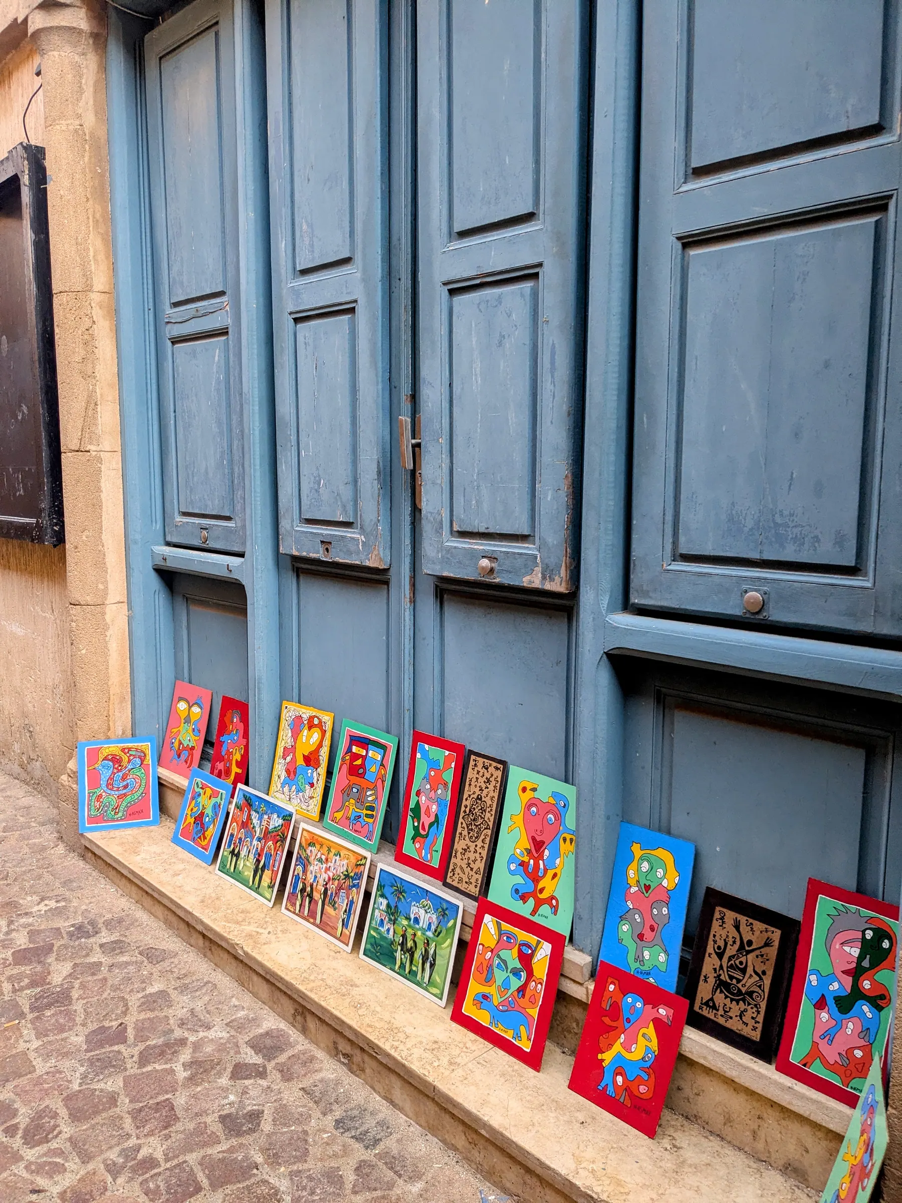 Paintings displayed below blue shutters in the Essaouira medina