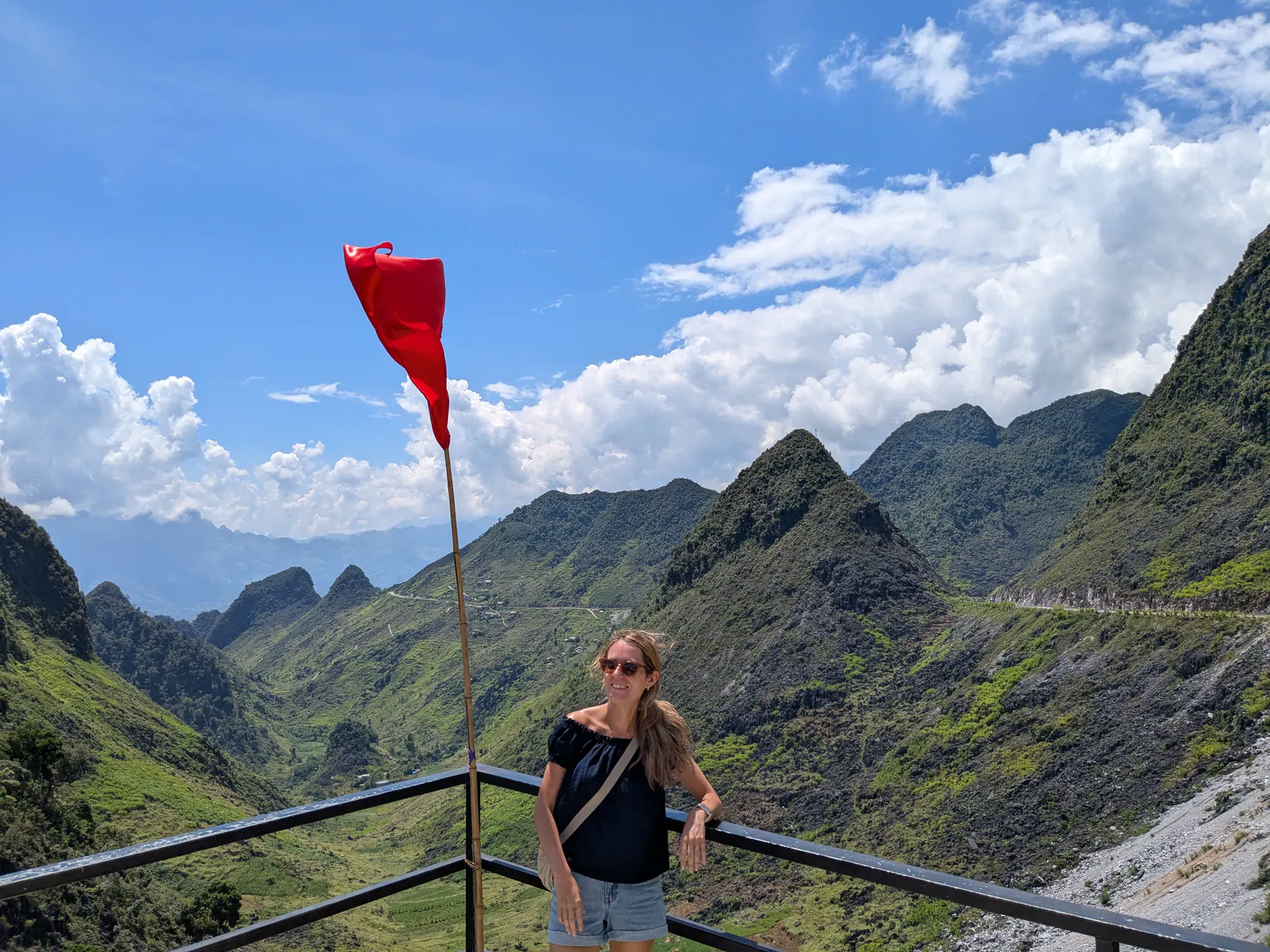 Zane standing by a cliffside railing beneath a Vietnamese flag overlooking layered mountains on the Ha Giang Loop