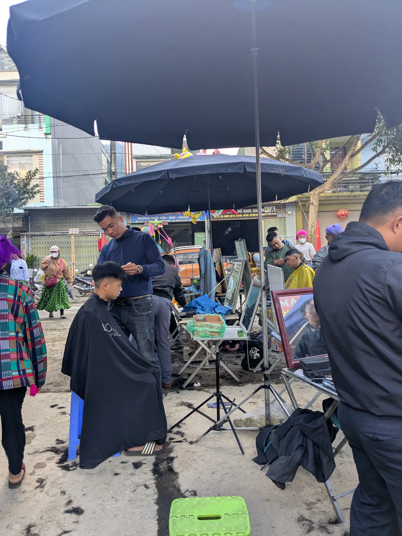 A barber cutting a seated man's hair beneath a large umbrella at Dong Van Sunday market