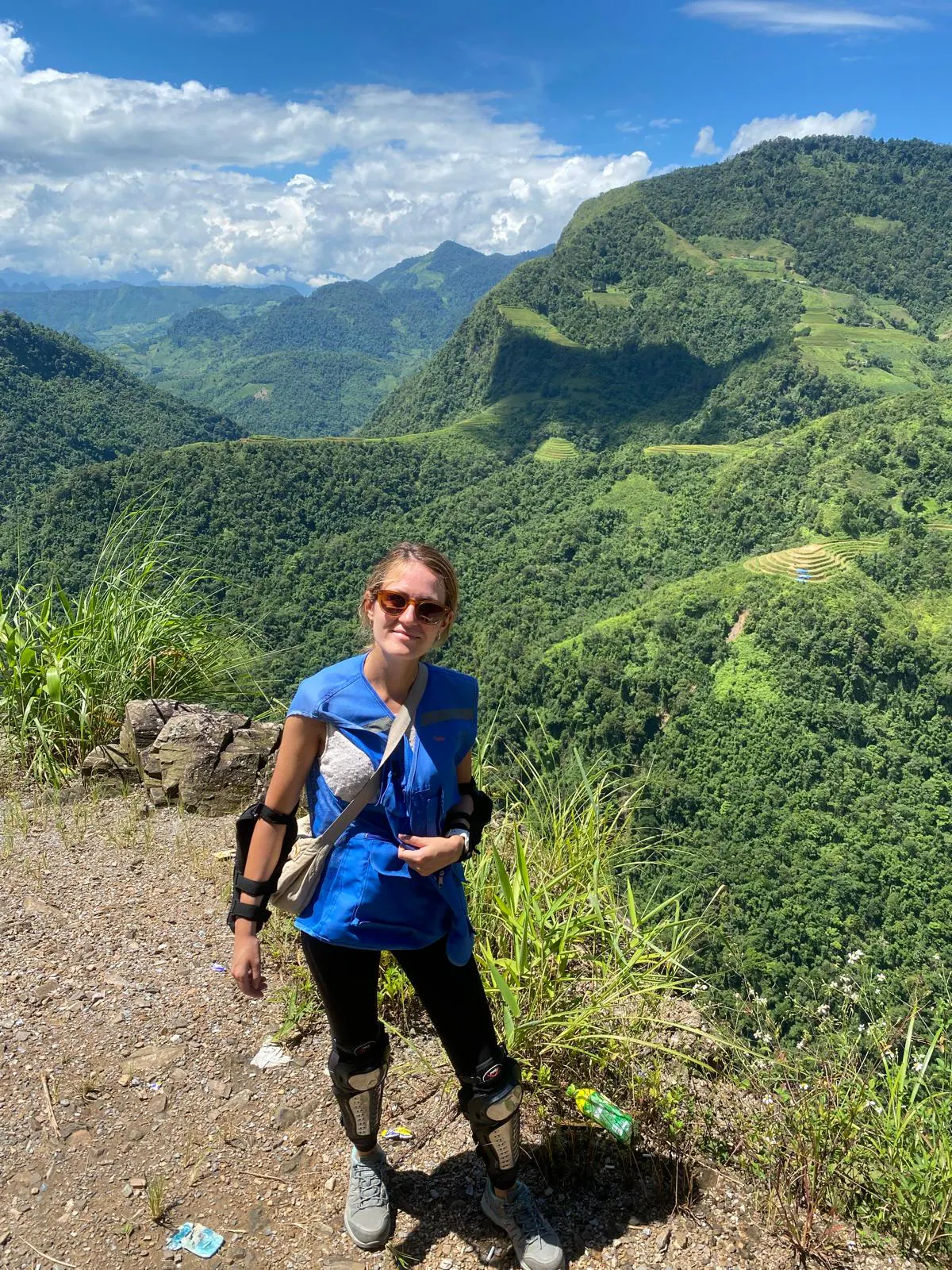 A traveller in a blue shirt wearing knee and elbow pads beside a green mountain valley on the Ha Giang loop