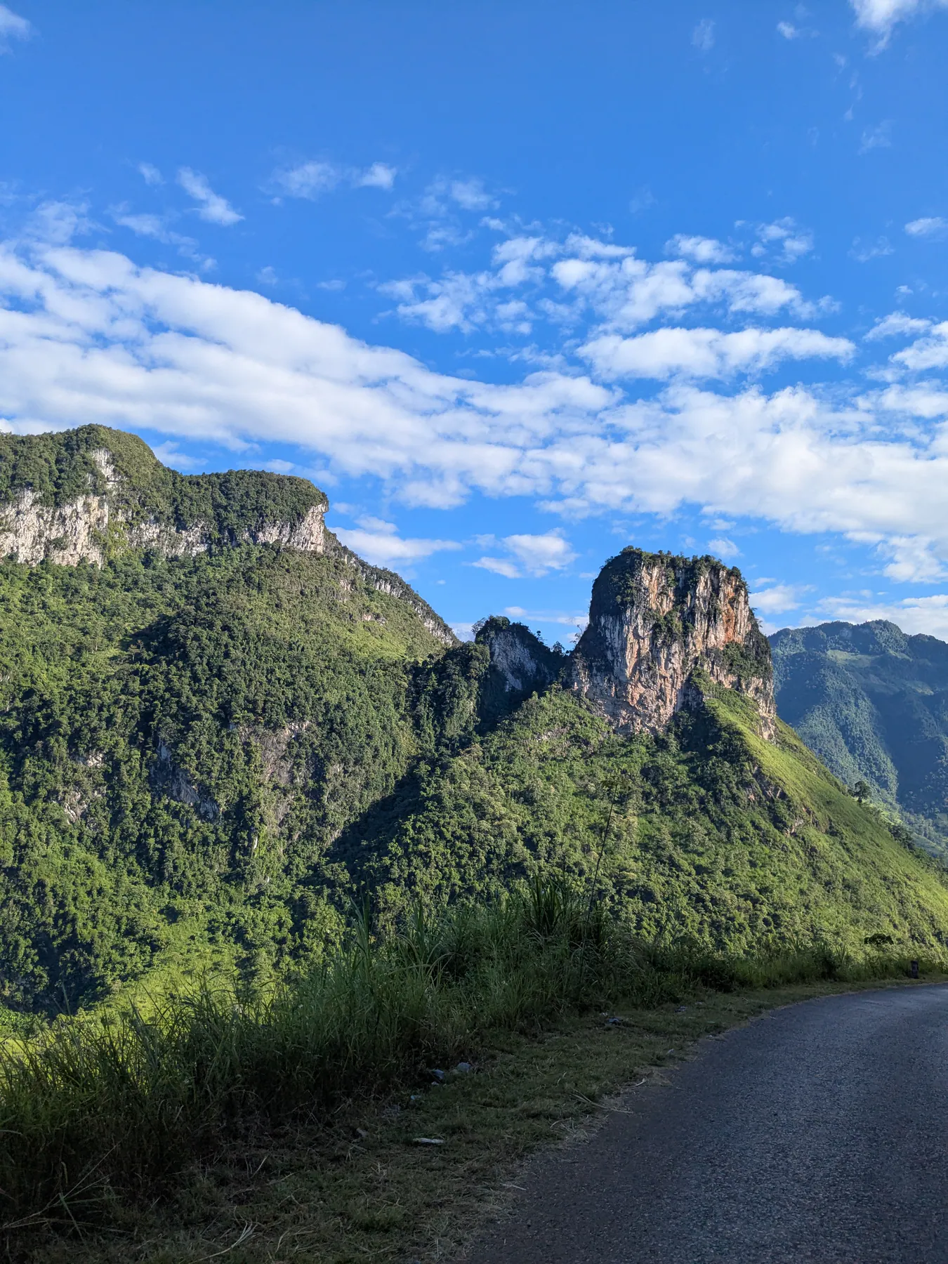 A paved mountain road curving towards tall sunlit limestone cliffs in Ha Giang