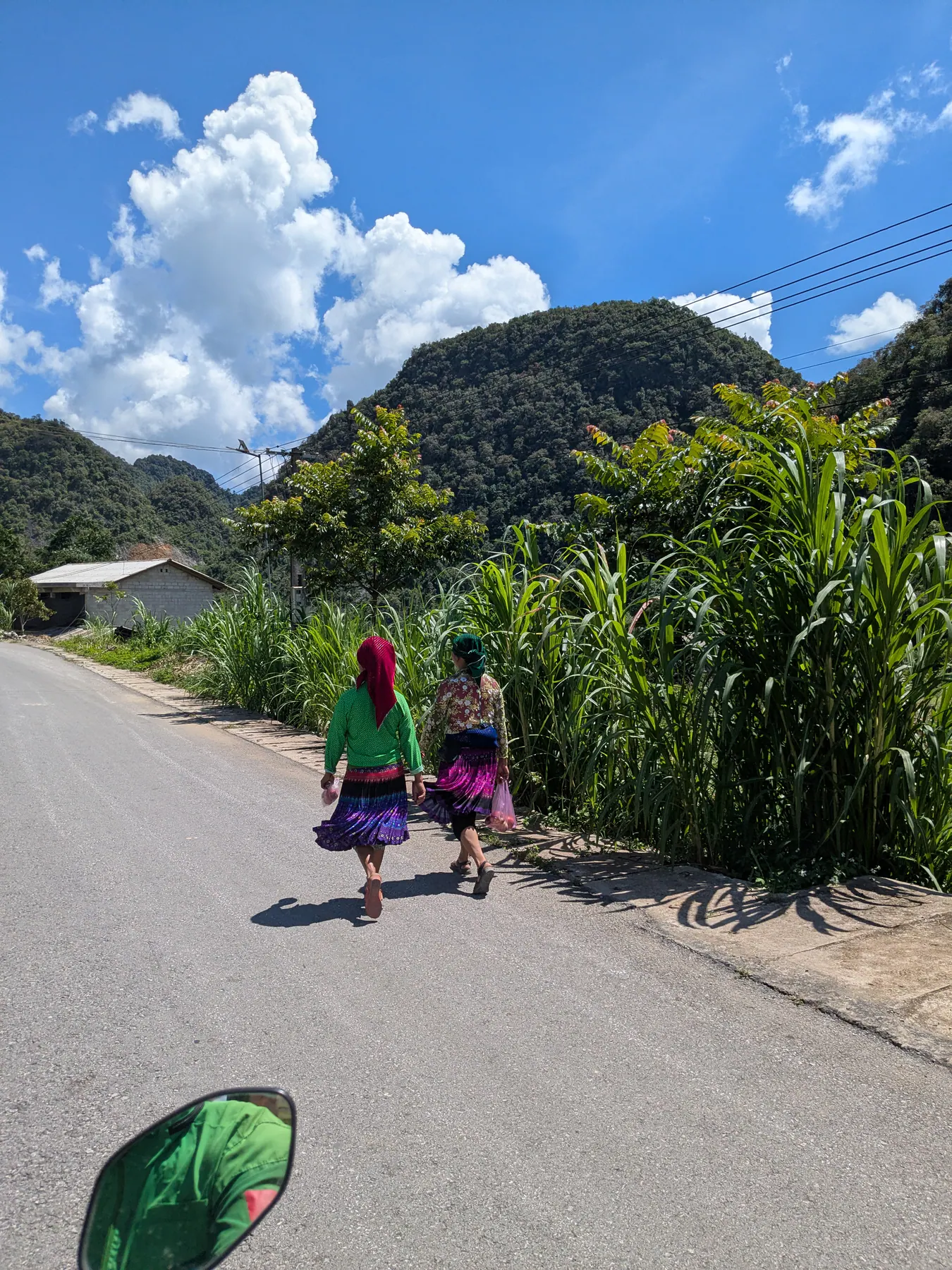 Two women in colourful clothing walking along a village road beside tall crops on the Ha Giang loop