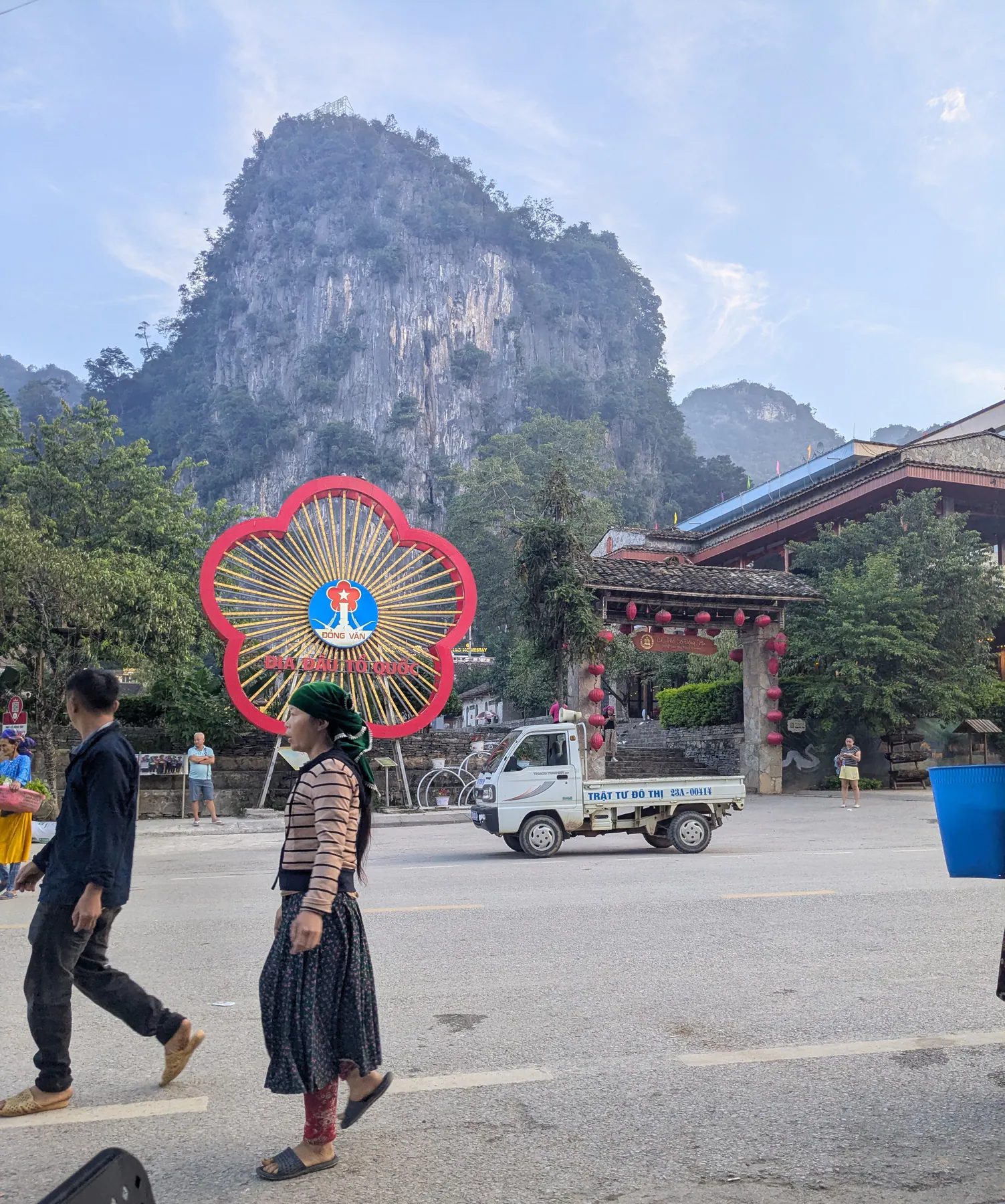Pedestrians passing a red flower-shaped Dong Van sign and a tiled gateway beneath a limestone mountain