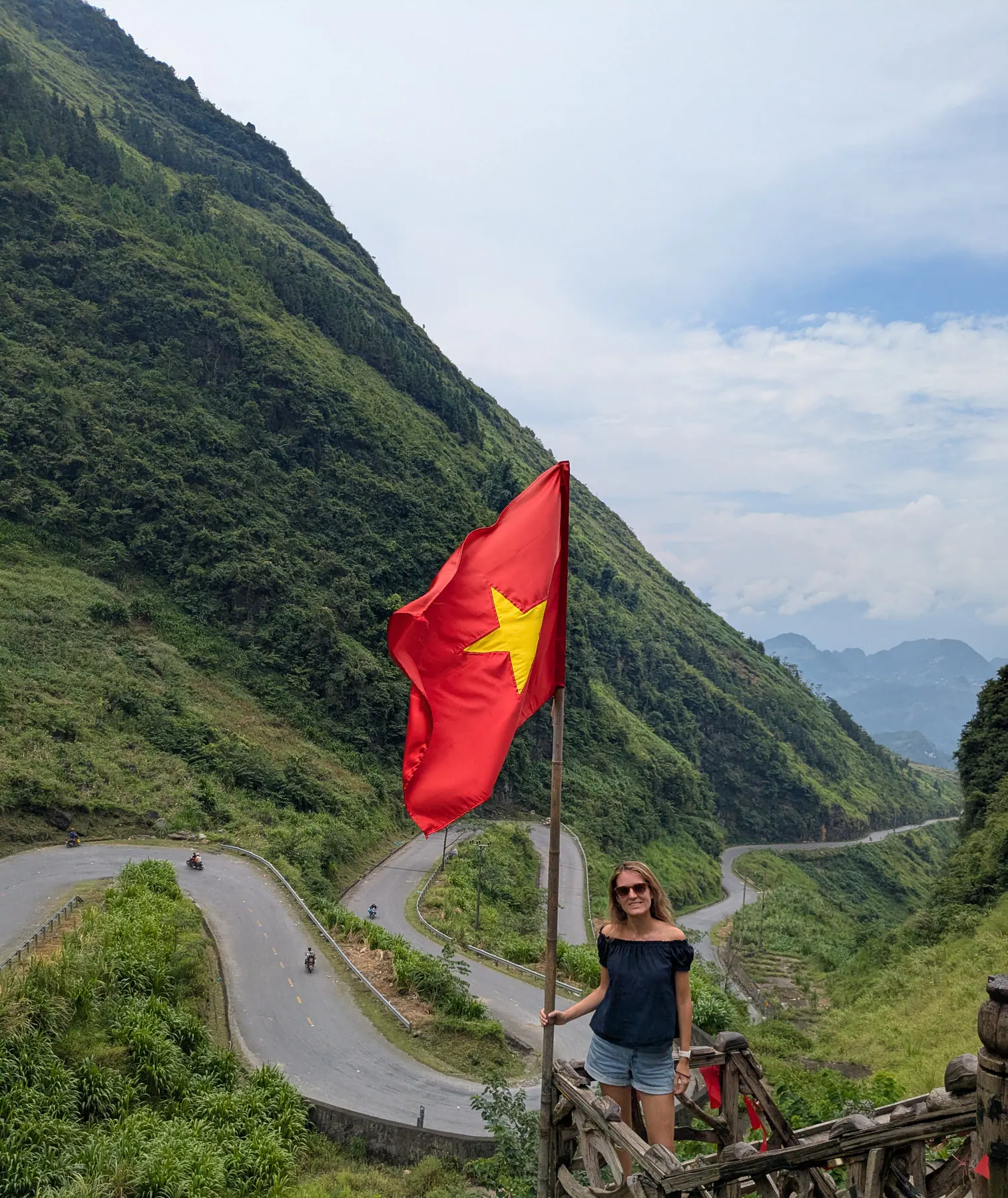Zane standing beneath a Vietnamese flag looking down over the tight hairpin serpentines of Tham Ma Pass in Ha Giang