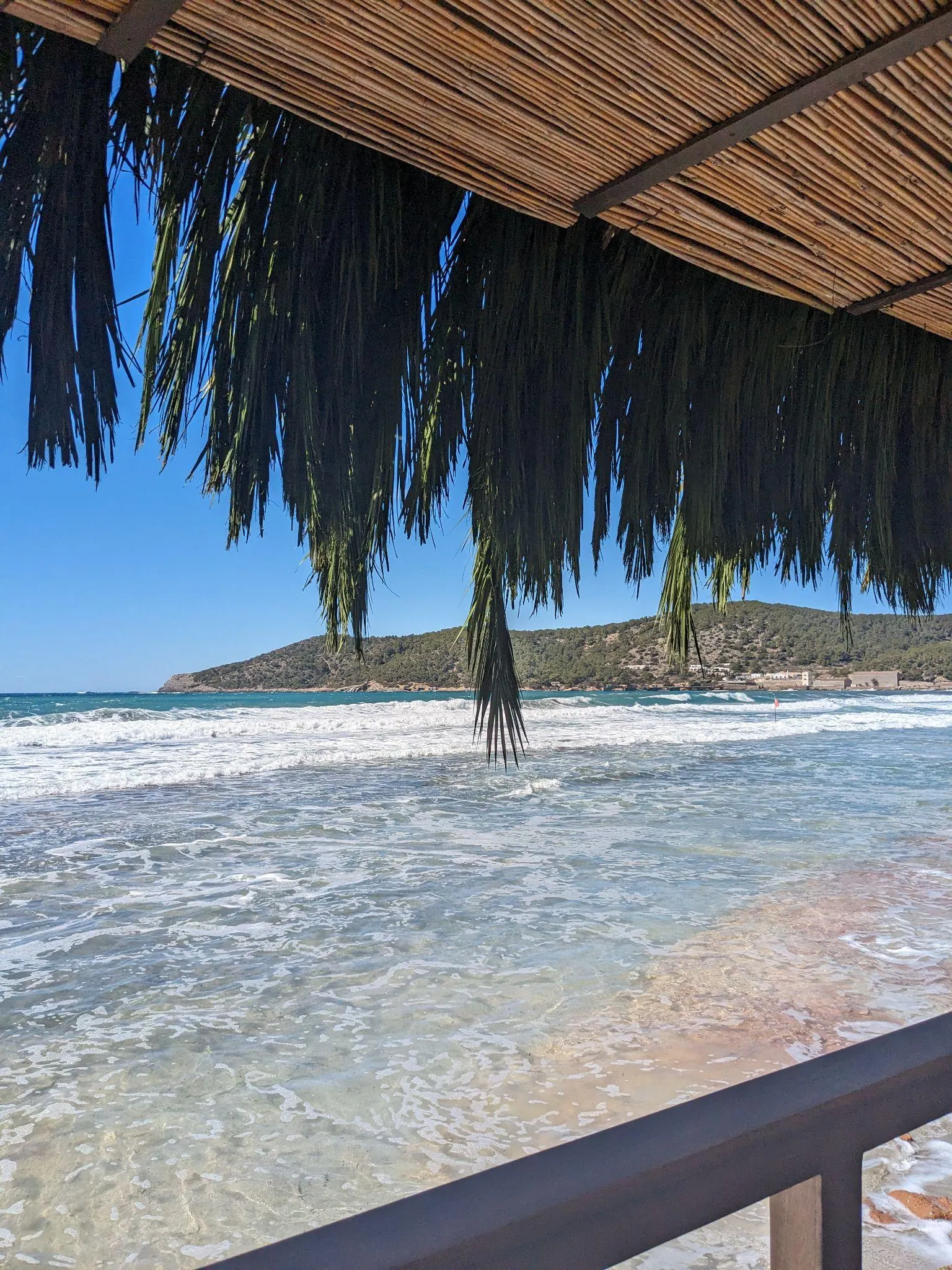 View of rolling waves and sea at Platja de Ses Salines from beneath a thatched beach shelter