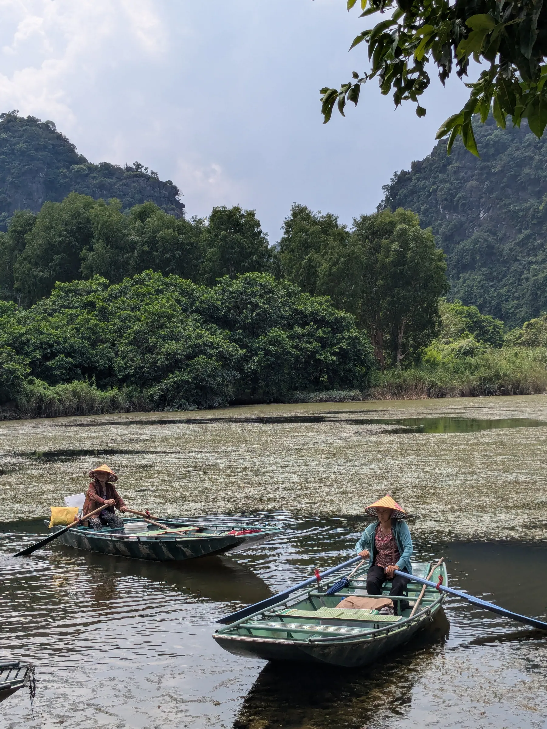 Two Vietnamese women wearing conical non la hats row traditional metal boats across calm water beneath limestone karst mountains in Ninh Binh