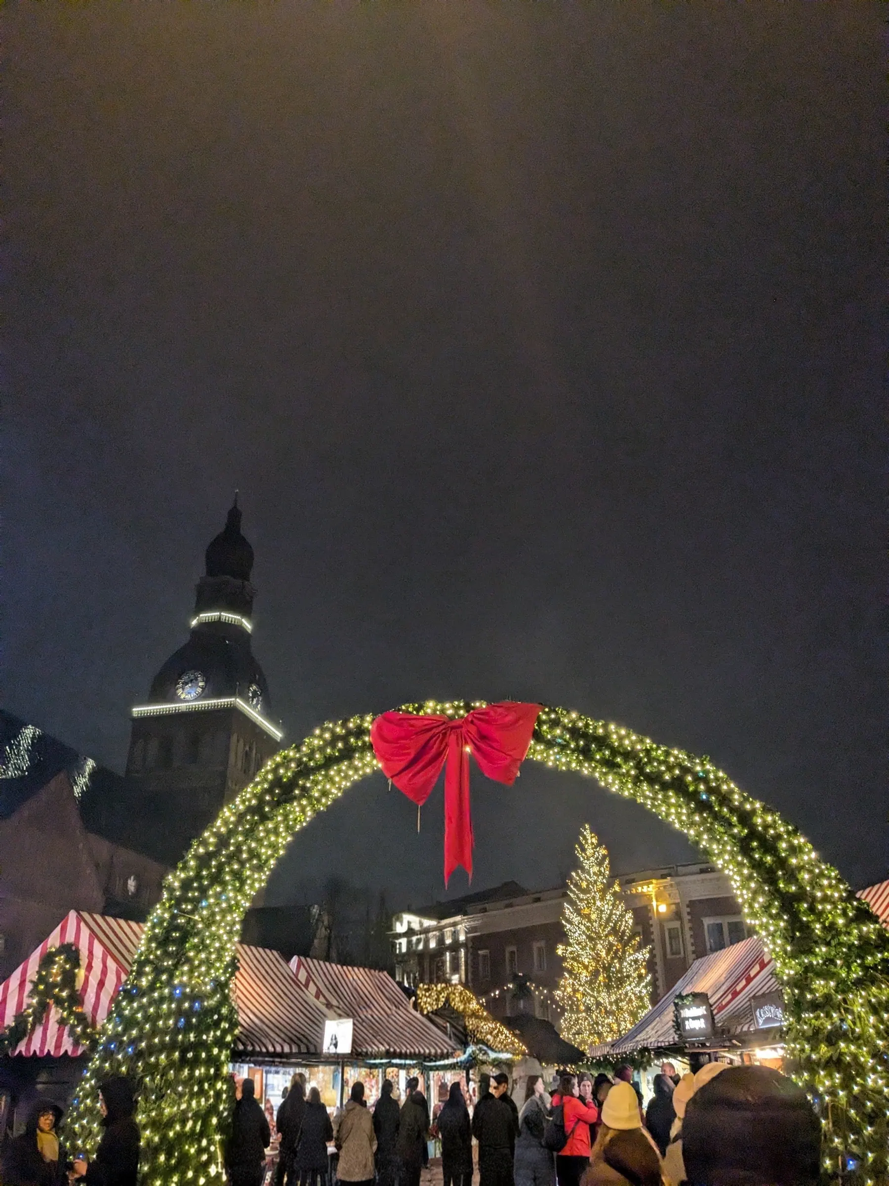 Arco iluminado con lazo rojo que enmarca el mercadillo navideño en la plaza de la Catedral y la torre de la catedral de Riga