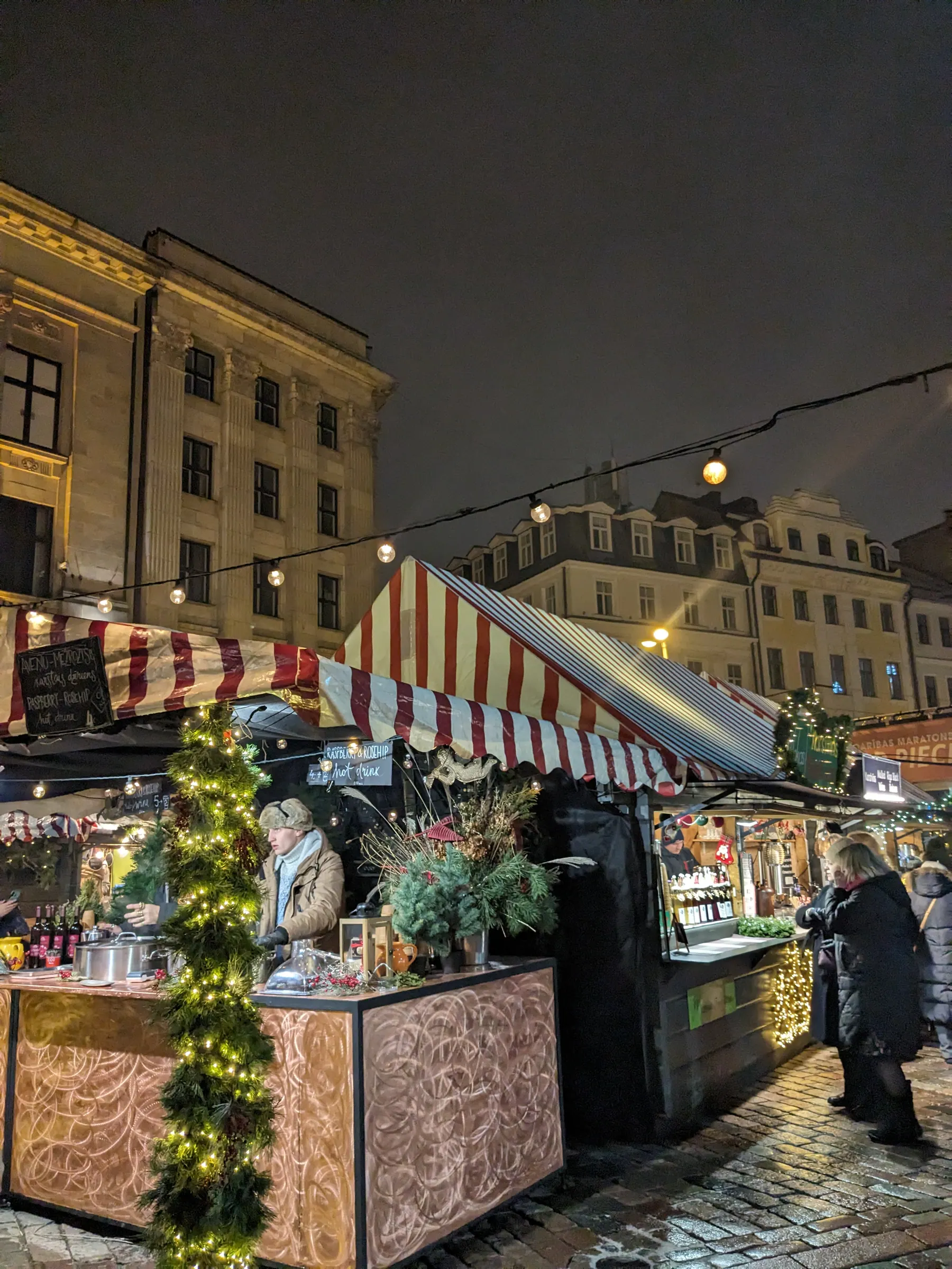 Festive wooden kiosk adorned with pine boughs serving steaming hot berry drinks and spiced karstvīns