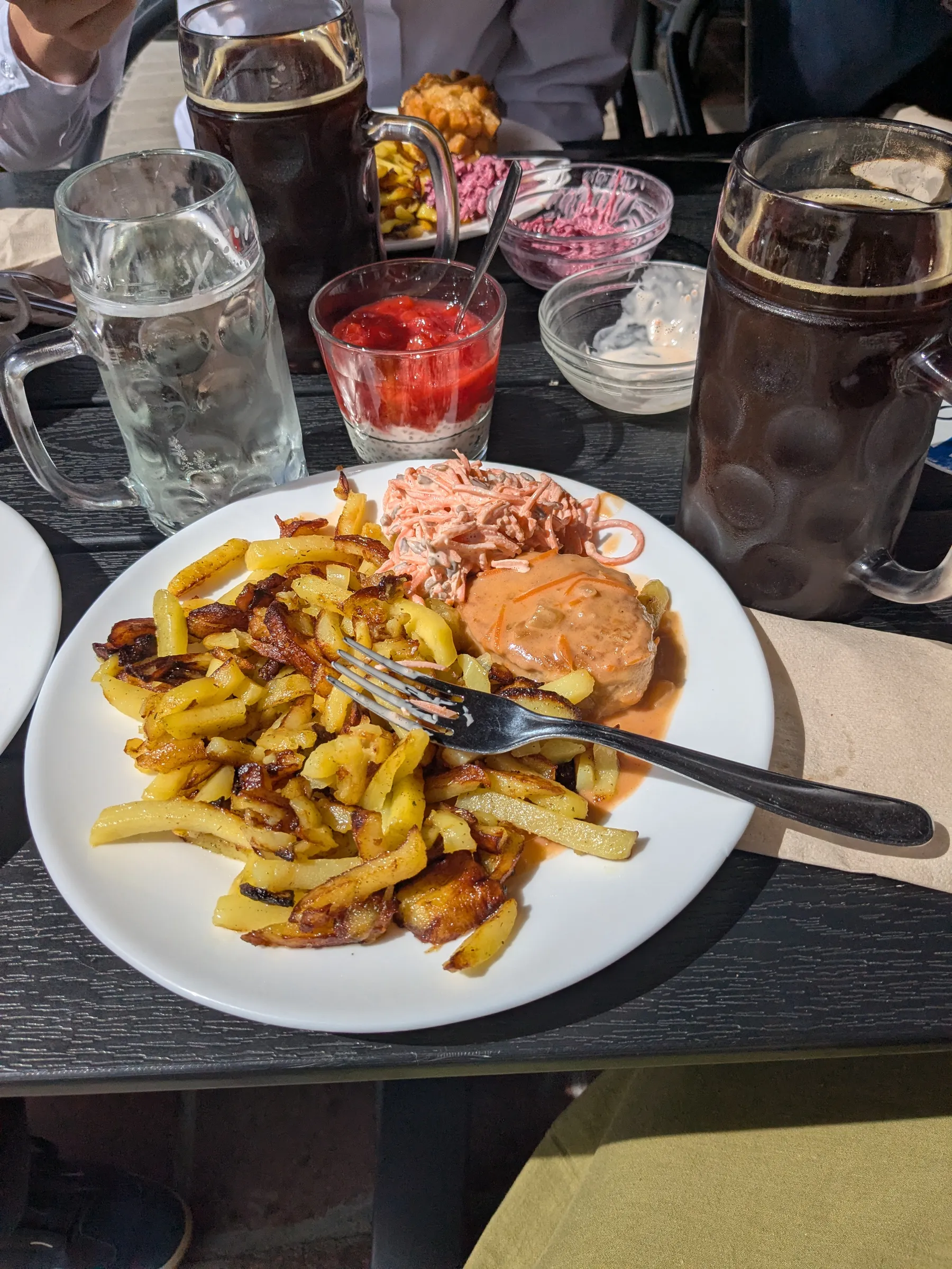 A traditional Latvian feast at Lido featuring golden fried potatoes, pork cutlet with sauce, fresh carrot salad, and cold dark kvass in dimpled beer mugs