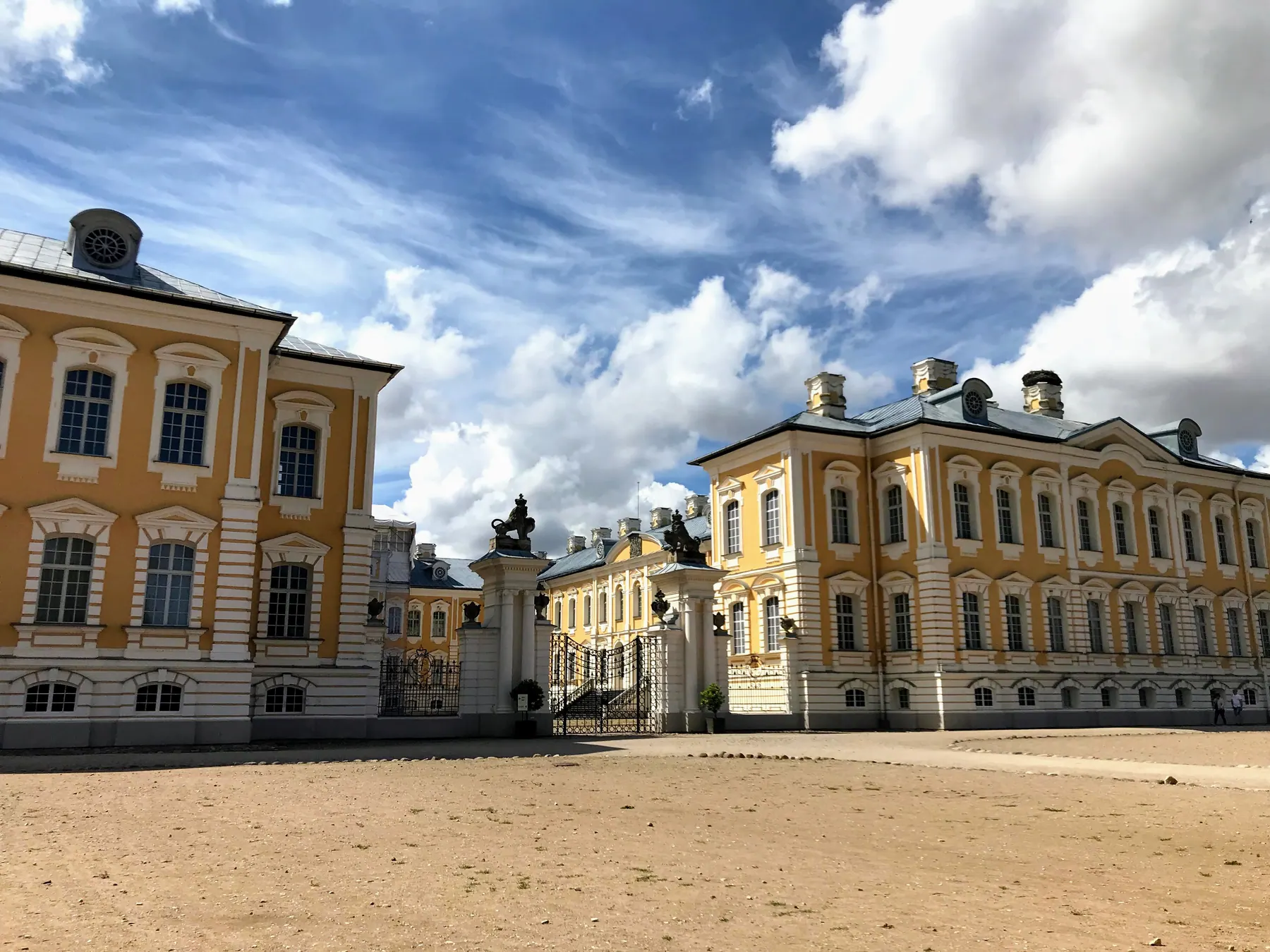 The yellow Baroque facade and ornate wrought iron entrance gates of Rundāle Palace beneath a summer sky in Pilsrundāle, Latvia