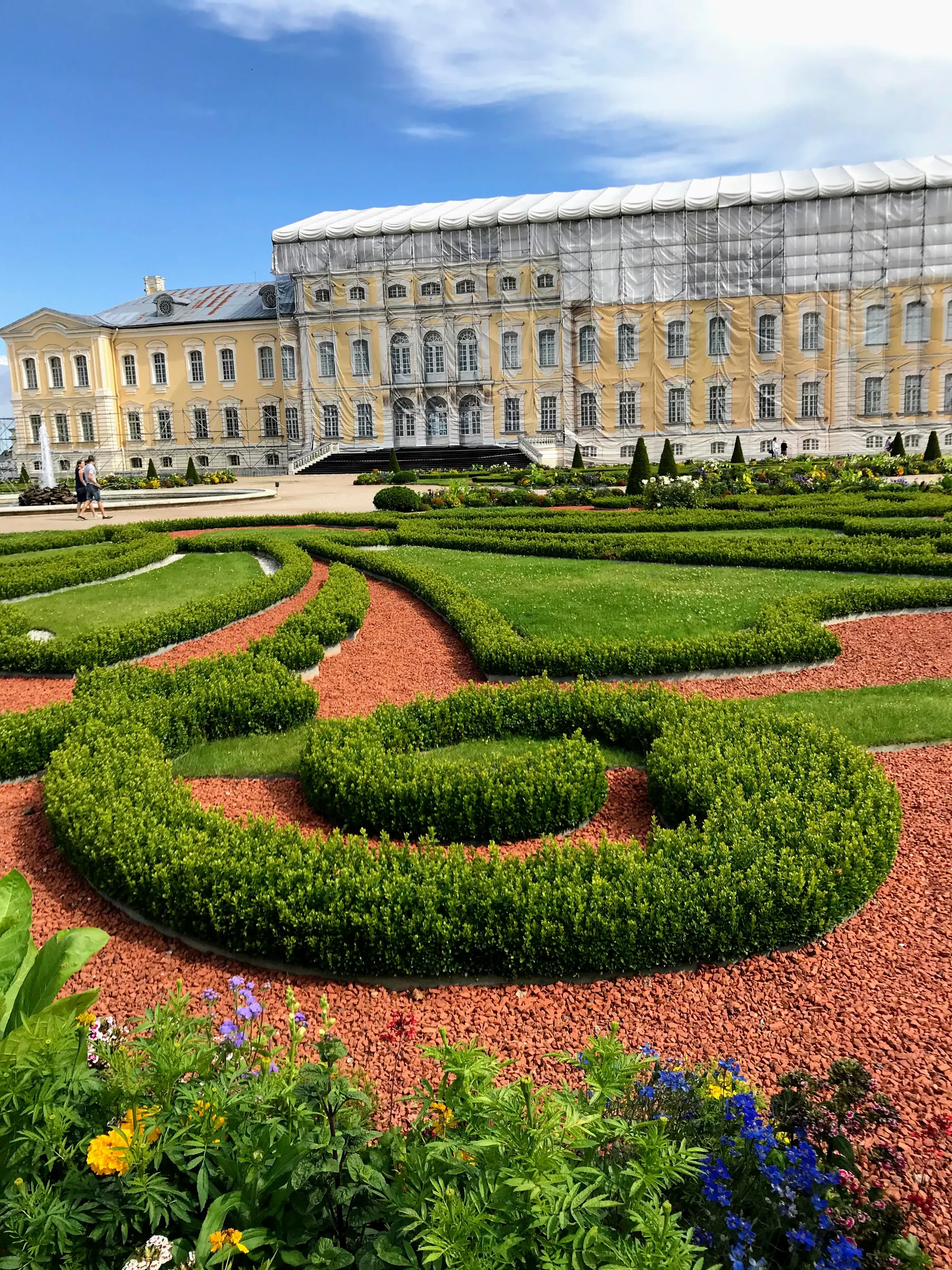 Symmetrical French garden parterres with clipped boxwood borders, colorful blooming flowerbeds, and the Baroque facade of Rundāle Palace in the background