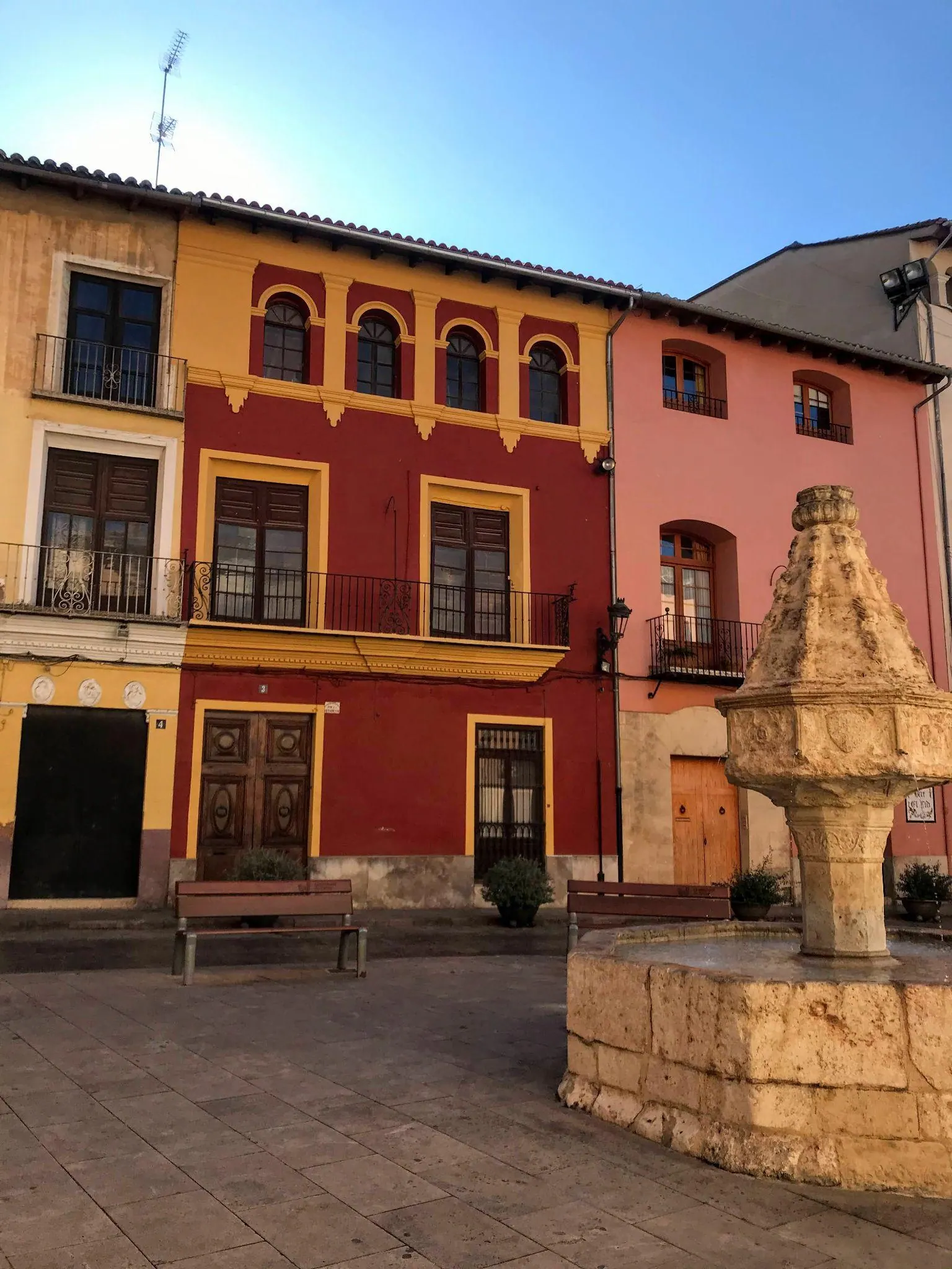 A peaceful historic square in Xàtiva with leafy trees, a stone fountain, and traditional townhouses
